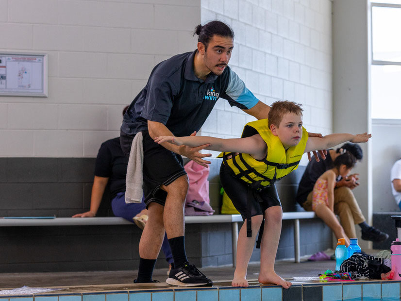 Child wearing a lifejacket practises survival skills at the side of the pool with a swim teacher