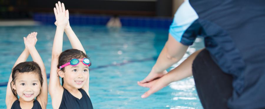 Two kids are learning hand movements with their swim instructor in front of the swimming pool.